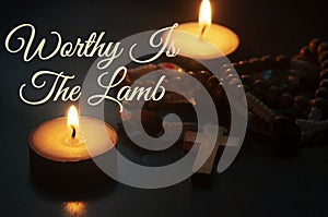 Worthy Is The Lamb text with Holy Rosary and burning candles background. Christian prayer and religion concept