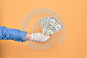 Wounded hand of caucasian young woman cover with bandage holding bunch of dollars banknotes over isolated yellow background