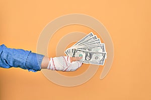 Wounded hand of caucasian young woman cover with bandage holding bunch of dollars banknotes over isolated yellow background