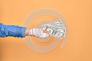 Wounded hand of caucasian young woman cover with bandage holding bunch of dollars banknotes over isolated yellow background