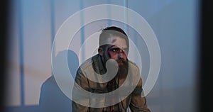 A wounded man with a beard sits behind a prison grate