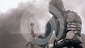 Wounded woman knight sits in smoke and leans on sword, slow motion