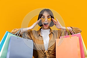 Wow, big sale. Emotional young woman wearing and touching sunglasses holding colorful shopping bags, yellow background