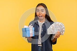 Wow, crazy gift shopping! Portrait of surprised girl in denim shirt holding present box, dollar money banknotes