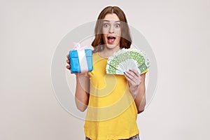 Wow, crazy gift shopping. Surprised girl in yellow casual T-shirt holding present box, euro money