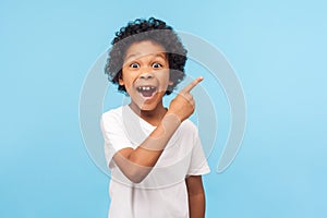 Wow look, advertise here! Portrait of amazed cute little boy with curly hair pointing to empty place