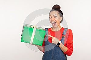 Wow, look at awesome gift! Portrait of joyful surprised girl with hair bun in denim overalls pointing at wrapped present box photo