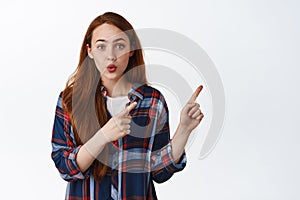 Wow look here. Amused millennial redhead girl, pointing right and pucker lips, say wow, showing advertisement, promotion