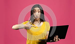 Wow offer concept. Portrait of shocked chinese lady holding laptop computer and pointing at it, pink background