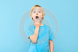 Wow! Portrait of a shocked cute little boy with blond hair covering open mouth with one hand, on pastel blue background.