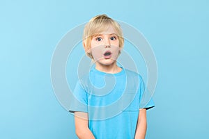 Wow! Portrait of a shocked cute little boy with blond hair on pastel blue background.