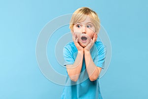 Wow! Portrait of a shocked cute little boy with blond hair on pastel blue background. Surprised preschooler studio shot.