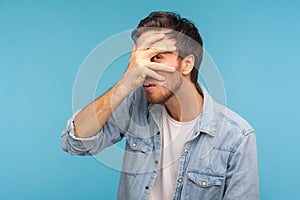 Wow, what is it? Portrait of curious nosy man in worker denim shirt looking through fingers with surprised