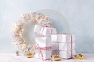 Wrapped boxes with presents,  decoraative golden cones, wreath with fairy lights  and decorative white tree on white marble agains