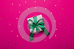 Wrapped present or giftbox with green ribbon on deep red or magenta background with hearts confetti, copy space, top view, flat