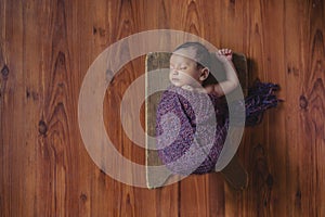 Wrapped in purple woolen cloth Indian 1 month baby boy sleeping on wooden board on wooden floor one hand up, newborn