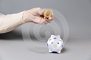 Wrinkled hand of an elderly woman holding a bitcoin near a piggy bank. Gray background. The concept of savings and