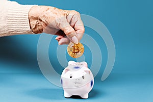 Wrinkled hand of an elderly woman puts a bitcoin in a piggy bank. Blue background. The concept of investment and