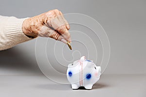 Wrinkled hand of an elderly woman puts a euro coin in a piggy bank. Gray background. The concept of savings and banking