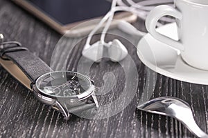 Wrist watch and mobile phone with headphones and a cup of coffee on a dark wooden table