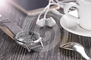 Wrist watch and mobile phone with headphones and a cup of coffee on a dark wooden table