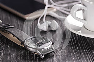 Wrist watch and mobile phone with headphones and a cup of coffee on a dark wooden table