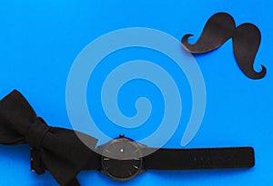 Wristwatch, paper mustache and bow tie on a blue background. Men`s black accessories. International Father`s Day.