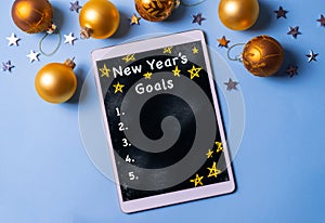 Writing new year`s goals list on a tablet on blue background with christmas golden balls and silver stars