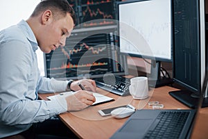 Writing on the notepad. Man working online in the office with multiple computer screens in index charts