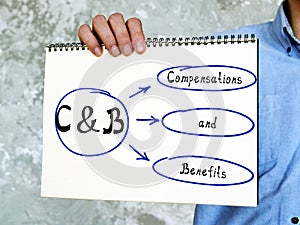 Written phrase C&B Compensations and Benefits . man`s hand holding hardcover kraft notebook