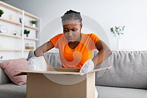Wrong parcel. Frustrated Afro woman unpacking carton box, unhappy with bad delivery service, sitting on couch at home