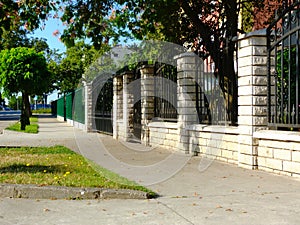 wrought iron fence between square stone piers and solid stone base. sidewalk with grass patch