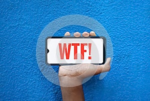 WTF wording on smart phone screen  on blue background with copy space for text. Person holding mobile on his hand and