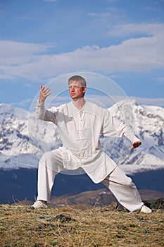Wushu master in a white sports uniform training kungfu in nature on background of snowy mountains