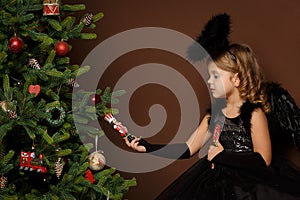 X-mas, winter vacation and people concept - little girl in black angel costume sits on a trunk near a Christmas tree and looks at