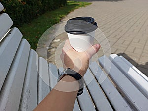 A& x27;s hand holds a white paper cup of coffee. Against the backdrop of a bench and nature. Black watch on hand