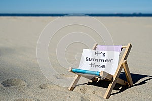 IT'S SUMMERTIME text on paper greeting card on background of beach chair lounge summer vacation decor. Sandy beach