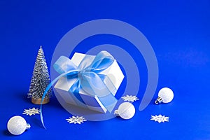 Xmas background blue. White gift box with blue ribbon, winter tree, Snowflakes and Silver balls in Christmas composition on blue