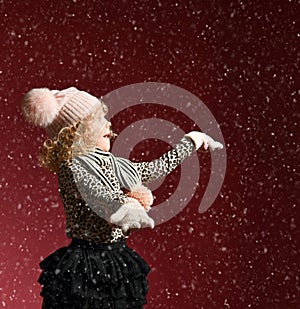 Xmas and New Year concept. Surprised baby girl in warm hat and gloves standing under snow, catching it on her open palms