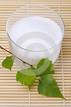 Xylitol birch sugar in glass bowl with birch leaves on bamboo mat