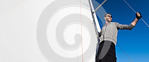 Yacht captain with a beard stands on sail boom on a sailing yacht, holding the rope in his hand and smiling, feeling