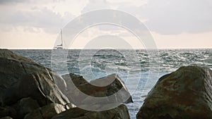 Yacht ship sailing at stunning sea view at sunset, waves break against big rocks and splash. Breathtaking ocean sunset