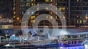 Yachts and boats with tourists staying near shoping mall and passing under a bridge in Dubai Marina district night