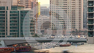 Yachts and boats with tourists staying near shoping mall and passing under a bridge in Dubai Marina district timelapse.