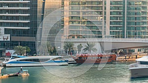 Yachts and boats with tourists staying near shoping mall and passing under a bridge in Dubai Marina district timelapse.