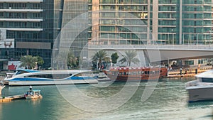 Yachts and boats with tourists staying near shoping mall and passing under a bridge in Dubai Marina district timelapse.