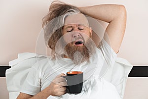 Yawning bearded man drinking morning coffee lying in bed. Sleepy guy trying to wake up. Closeup