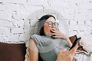 Yawning sleepy caucasian woman in eyeglasses using mobile phone while resting on a sofa at home