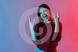 Yeah, that`s crazy! Neon light portrait of delighted happy woman showing rock and roll hand gesture, punk sign