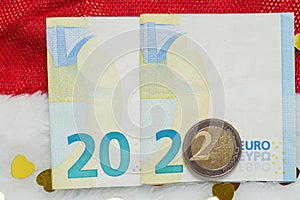 Year 2022 made out of Euro bank notes and coin. On red Santa hat and golden heart shape confetti. New year celebration and change
